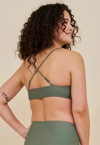 Reggiseno sportivo verde con una texture liscia, caratterizzato da spalline incrociate regolabili e hardware metallico, su uno sfondo neutro.