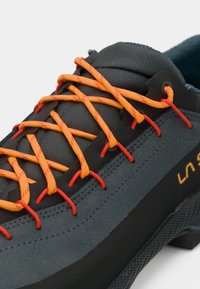 Sapatilha de escalada cinza com parte superior texturizada, atacadores laranja e vermelho, biqueira em borracha e sola robusta para aderência. Logótipo da marca visível ao lado.