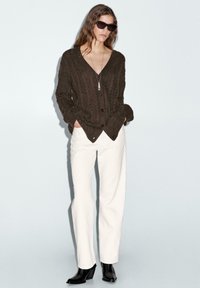Bruine gebreide cardigan met een V-hals en knopen, gecombineerd met witte hoge taille wijde broek en zwarte enkellaarsjes.