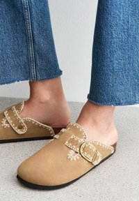 Suede slip-on clogs in light tan with decorative stitching and two buckles. Black rubber sole. Worn with blue denim flared pants.