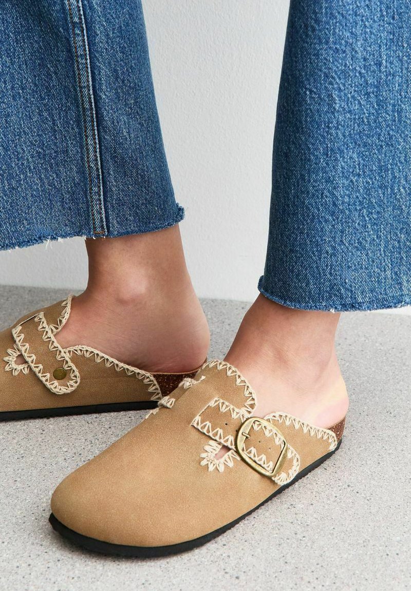 Suede slip-on clogs in light tan with decorative stitching and two buckles. Black rubber sole. Worn with blue denim flared pants.