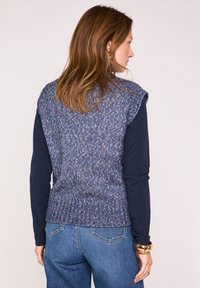 Gilet en tricot dans des tons de bleu avec une texture côtelée ; associé à une chemise à manches longues navy. Présente des coutures visibles et des détails aux épaules.
