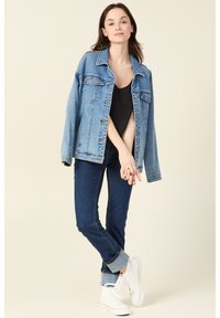 Hellblaues Denim-Jacke mit lässiger Passform, ausgestattet mit zwei Brusttaschen, Knopfverschluss und fransigen Kanten. Kombiniert mit dunklen Jeans und weißen Sneakers.