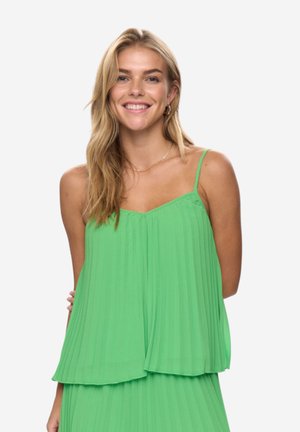 Usměvavá žena s dlouhými blond vlasy, oděná v zeleném plisovaném topu na úzká ramínka a odpovídající sukni, stojící na bílém pozadí.