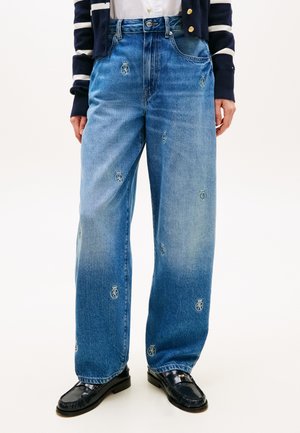 Blå jeans med vide ben og små broderede kroner og skjoldlogoer, båret med sorte loafers og en marineblå stribet cardigan.