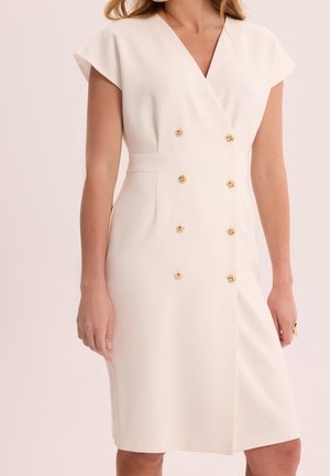 Femme portant une robe blanche mi-longue avec des manches courtes et six boutons dorés en deux rangées verticales sur le devant.