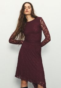 Bordeauxrotes Spitzenkleid mit langen Ärmeln, strukturiertem Stoff, figurbetontem Oberteil und asymmetrischem Saum; kunstvolles Blumenmuster auf den Ärmeln.