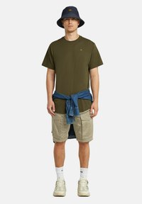 Camiseta de manga corta de color verde oliva, pantalones cortos de carga beige con bolsillos, camisa de denim atada a la cintura, sombrero de cubo azul, zapatillas beige.