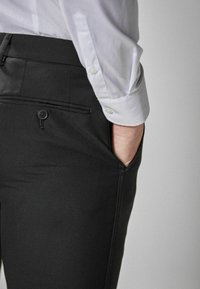 Čierne nohavice z hladkého materiálu s bočným vreckom a zadným vreckom na gombík. Čiastočne viditeľná biela košeľa.