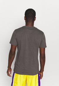 Hombre con una camiseta de manga corta gris oscuro y pantalones cortos amarillos brillantes con rayas moradas, de espaldas contra un fondo blanco liso.
