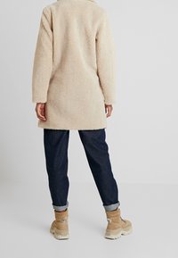 Abrigo largo beige y esponjoso con cuello y mangas largas, combinado con vaqueros azul oscuro remangados y botines beige.