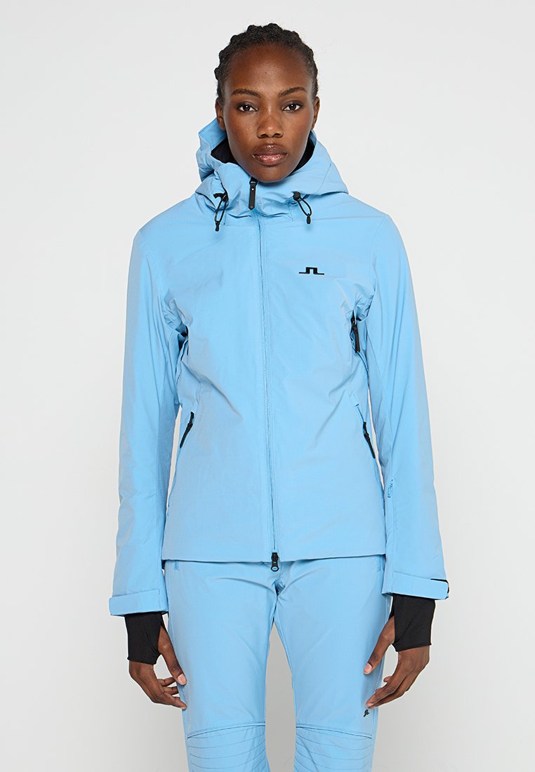 Kobieta w jasnoniebieskiej kurtce z kapturem na zamek błyskawiczny i dopasowanych spodniach, stojąca na tle jednolitego białego tła.