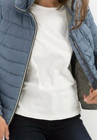 Polstret blå jakke med quiltet mønster, uldret for at afsløre en grå foring, båret over en ensfarvet hvid skjorte, parret med mørke jeans.