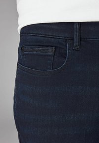 Jeans de mezclilla azul oscuro con una textura suave, que presentan un bolsillo frontal, trabillas para el cinturón y costuras en contraste. Vista de cerca del lado.