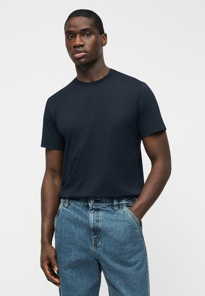 Άνδρας που φοράει ένα απλό σκουρογάλανο μπλουζάκι τυλιγμένο μέσα σε ανοιχτό μπλε τζιν παντελόνι, με το ένα χέρι στην τσέπη, στέκεται μπροστά σε μια απλή ανοιχτή φωτεινή επιφάνεια.