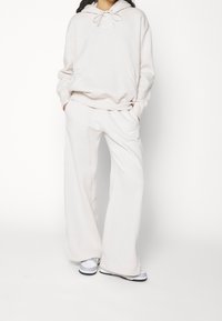 Béžová mikina Nike s kapucí, se stahovacími šňůrkami a klokaní kapsou, doplněná o široké tepláky se stejným motivem a logem, nošená s teniskami.