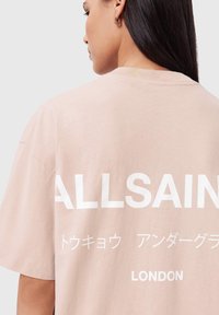 Lichtroze oversized T-shirt met witte tekst 'ALLSAINTS' en 'LONDON'. Voorzien van korte mouwen en een ronde hals. Zachte stoftextuur.