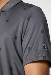 Polo gris avec une texture lisse, présentant un motif subtil de logos noirs. Comprend un accent de logo sur la manche.