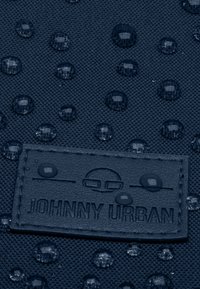 Hajószerű textúrájú anyag vízcseppekkel, amelyen a "JOHNNY URBAN" felirattal ellátott bőr címke található. Kör- és ovális cseppek láthatók.