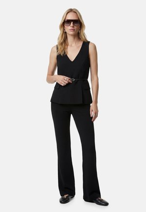 Nainen, joka pukeutunut mustaan hihattomaan V-kaulaiseen toppiin, jossa on vyö, mustiin housuihin, mustiin loafereihin, aurinkolasit ja hopeasormukset vaaleaa taustaa vasten.