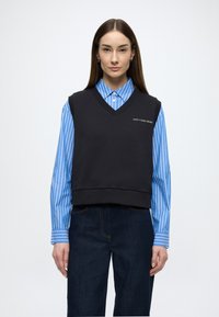 Camisola preta sem mangas com decote em V, com um logótipo; usada sobre uma camisa de gola com riscas azuis e brancas. Jeans de ganga escura completam o conjunto.