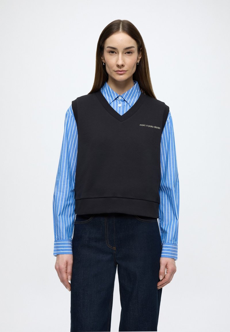 Camisola preta sem mangas com decote em V, com um logótipo; usada sobre uma camisa de gola com riscas azuis e brancas. Jeans de ganga escura completam o conjunto.