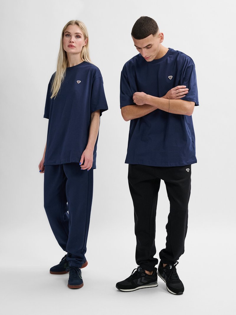Twee volwassenen dragen navyblauwe oversized t-shirts met kleine logo's, gecombineerd met navy sweatpants en zwarte joggers, staand tegen een effen achtergrond.