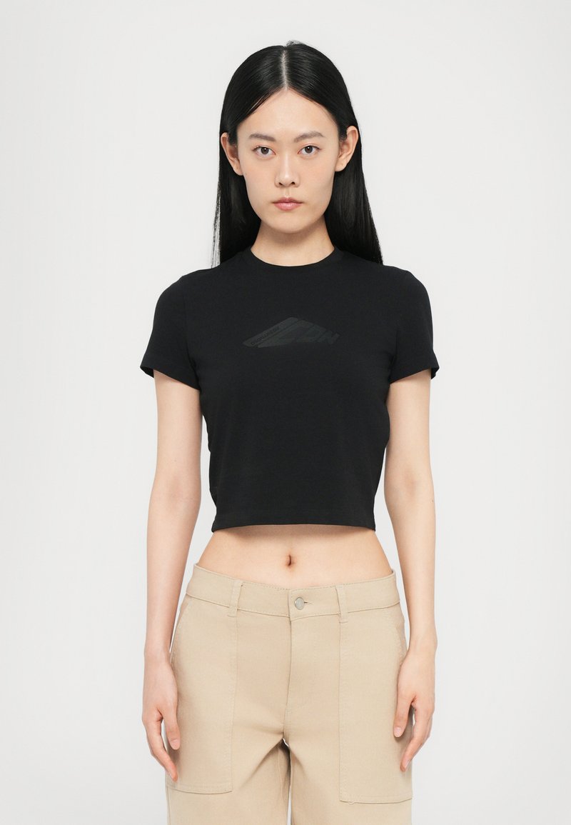 Black cropped t-shirt made of cotton with a round neckline. Features a subtle tonal graphic on the front. Worn with light beige pants.