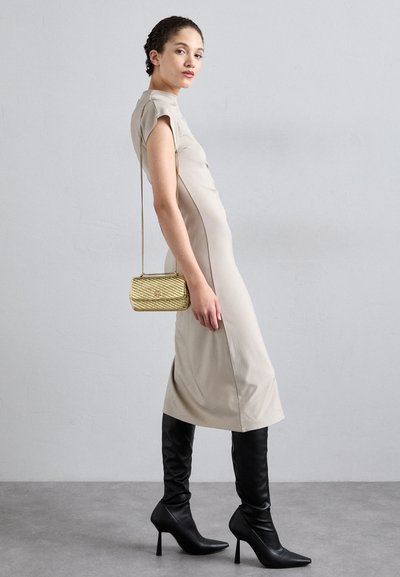 Vestido ajustado beige con mangas cortas, abertura lateral, combinado con botas negras de tacón alto hasta la rodilla y un pequeño bolso con cadena dorada.