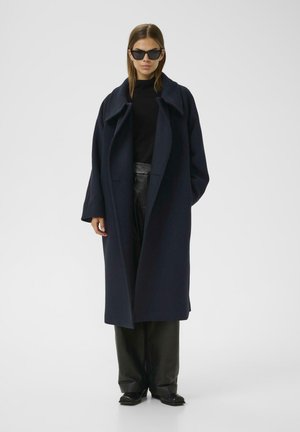 Navy wool overcoat with a loose fit, wide lapels, long sleeves, and side pockets. Worn over a black turtleneck and black leather pants.