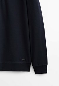 Sweat-shirt noir à manches longues avec poignets et bord côte, avec une petite étiquette rectangulaire de la marque près du bord inférieur.