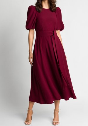 Femme portant une robe midi bordeaux avec des manches bouffantes et un lien à la taille, associée à des sandales à talons hauts avec bride à la cheville couleur nude.