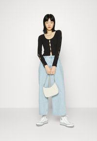 Camisola preta de mangas longas com recortes em forma de olho, combinada com jeans de perna larga azul-claro e ténis brancos de cano alto, segurando uma pequena mala branca.