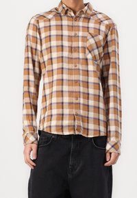 Man in een bruin, beige en blauwe geruite button-up shirt met borstzak, gecombineerd met losse zwarte broek, staand tegen een witte achtergrond.