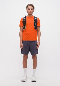 Νεαρός άνδρας με πορτοκαλί κοντομάνικο μπλουζάκι, μαύρο γιλέκο ενυδάτωσης με μπουκάλια νερού, γκρι σορτς, λευκές κάλτσες και λευκά παπούτσια τρεξίματος.