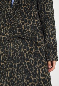 Manteau à imprimé léopard en vert foncé et beige, avec un tissu texturé, des fermetures à boutons et une poche latérale.