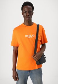 Oranžinė medvilninė marškinė su užrašu "REPLAY SINCE 1981" balta spalva, derinamos su mėlynomis džinsinėmis kelnėmis ir mažąja juoda per petį nešiojama rankine.