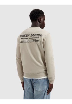 Man draagt beige sweatshirt met zwarte tekst 'BALLIN STUDIO Records & Publishing' op de achterkant, gecombineerd met lichtblauwe spijkerbroek, met zijn rug naar de camera toe.