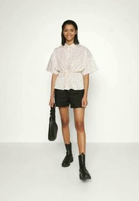 Blusa branca com padrão floral, mangas curtas, cintura ajustada, combinada com calções pretos e botas de tornozelo. Modelo segura uma bolsa preta.