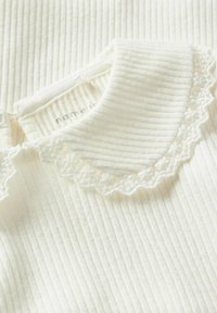 Weißer, gerippter Stoff mit einem mit Spitze verzierten Kragen. Besitzt eine weiche Textur und dezente vertikale Streifen, mit teilweise sichtbarem Etikett.