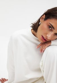 Witte sweatshirt met een ronde hals en een zachte textuur, gecombineerd met lichtgekleurde stof. Model met kort, donker haar en zilveren oorbellen.