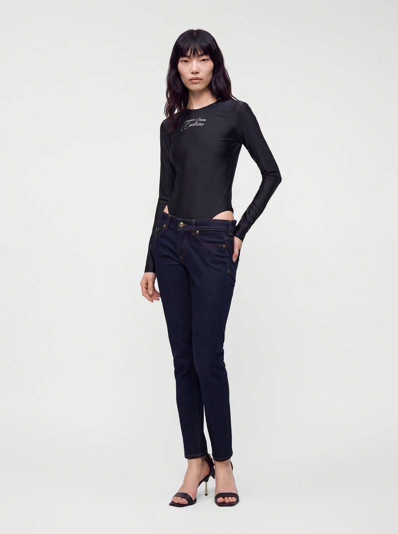 Femme aux longs cheveux noirs portant un haut noir ajusté à manches longues et un jean bleu foncé avec des sandales noires à talons hauts sur un fond uni.