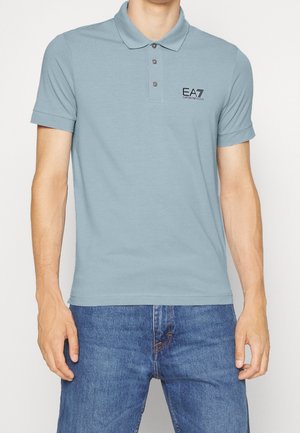 Světle modrá polokošile z bavlny s límečkem, třemi knoflíky a vyšívaným logem "EA7" na levé straně hrudi. Nošena s modrými džínami.