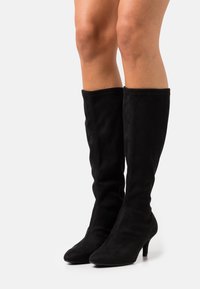 Beine, die schwarze kniehohe Wildlederstiefel mit spitzen Zehen und niedrigen Kitten-Heels auf weißem Hintergrund tragen.