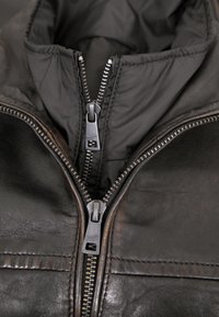 Schwarze Lederjacke mit hohem Kragen, ausgestattet mit zwei silbernen Reißverschlüssen und einem gesteppten Innenfutter. Strukturierte Oberfläche mit dezenten Nähten.