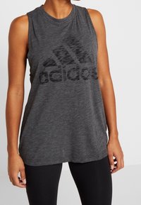 Šedý bezrukávový sportovní top vyrobený z měkké tkaniny, s výrazným černým logem Adidas přes hruď, s uvolněným střihem a kulatým výstřihem.