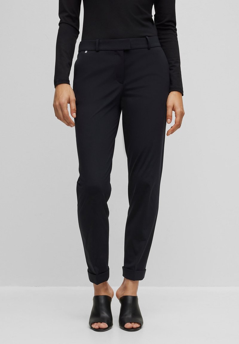 Pantalon noir sur mesure, coupe slim, avec une ceinture à passants et des revers retroussés, porté avec des mules noires à bouts ouverts.