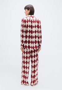 Femme debout de dos portant une chemise à manches longues et un pantalon jambes larges assortis, à motif losanges bordeaux et blanc, sur fond blanc.