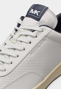 Hvid perforeret sneaker med hvide snørebånd, navy tungeetiket med "MK" og lys beige sålekant synlig.