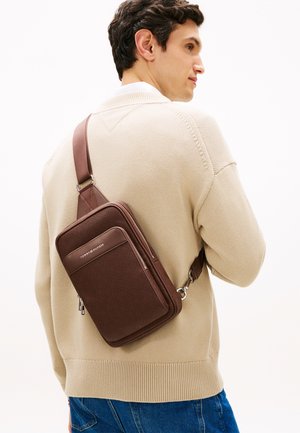 Καφέ υφασμάτινη τσάντα χιαστί από συνθετικό υλικό, με μπροστινή τσέπη με φερμουάρ και ρυθμιζόμενο λουράκι, φορεμένη πάνω από μπεζ πουλόβερ.